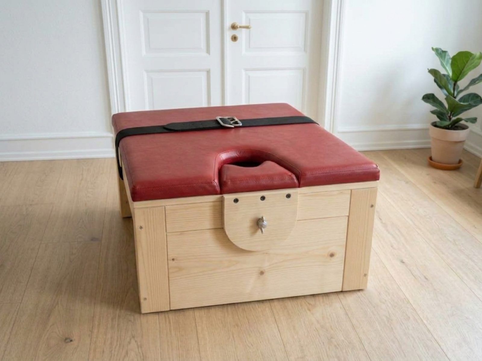 Pine facedown bench with red faux leather padded top, face hole and black buckled strap, wooden face rest held by a wing nut, in a white room with a plant